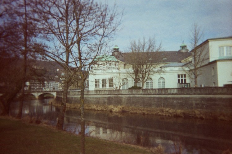 The view of the white-painted Kurhaus as seen from the other side of the river with some trees on this side and one thin bare one on the other side next to the Kurhaus.