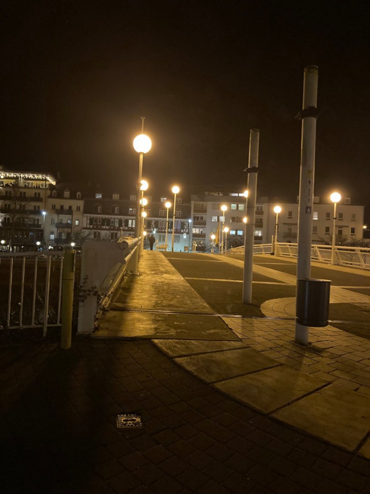 Bad Ems by night, a wide footbridge with strange pillars sticking out of it and buildings visible on the opposite side with no lights on in any of the windows.