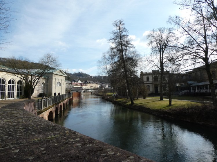 A view from a bridge in Bad Kissingen, looking down the river with the park on the right and the Wandelhalle on the left.