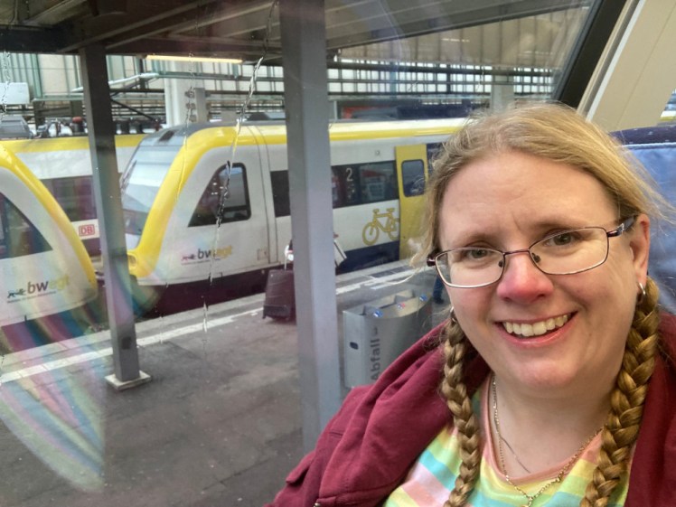 Me, grinning, in a pastel rainbow-striped top and dark red hoodie, on the top floor of a train at Stuttgart. The photo is angled so you can see the platform next to me is below me and that I'm at about roof-height of the single-decker train on the other side of the platform.