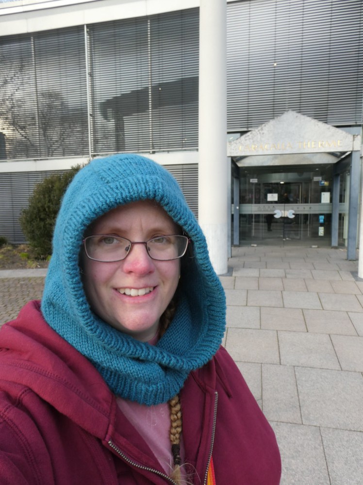 Me, standing outside the entrance in a blue knitted hood, dark red hoodie and pink t-shirt, with that modern/Classical facade behind me.