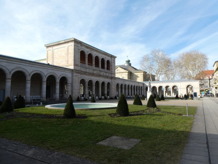 The Kurhaur, a stone one-storey buildig with arcades along the front. There is a small section of second-storey exactly matching the downstairs perched on top. In front are gardens, probably not looking their best at this time of year.
