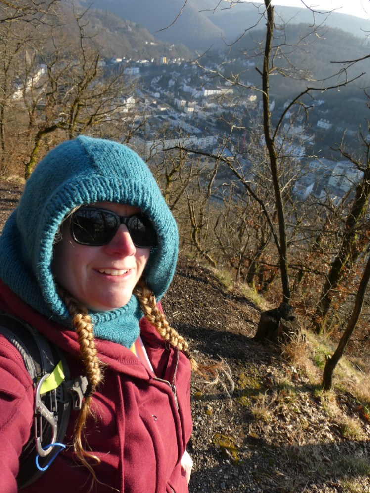 A selfie on the edge of the trail that kind of gives a view through the leafless trees to the town below. I'm wearing sunglasses, my blue knitted hood and my red hoodie.