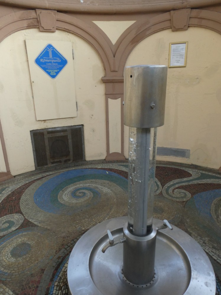 A pair of taps coming out of a clear pillar with bubbling water in it. The tap and pillar are in a little well in the ground with red and blue spiral tiled mosaics on the floor.