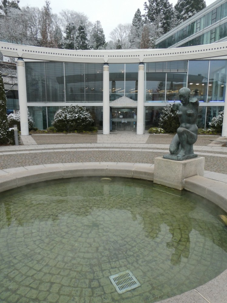 Right outside the Therme is a small shallow pool of greenish water, steaming, with a statue looming over it seen on the right in this picture with the modern/Classical building behind it.