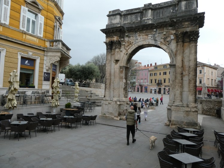 The Arch of the Sergii, a Roman arch standing in the narrow street with cafes on both sides, a square and an ordinary road behind it. This is the more ornate side of the arch but it's so blackened with age that it's hard to make out the details.