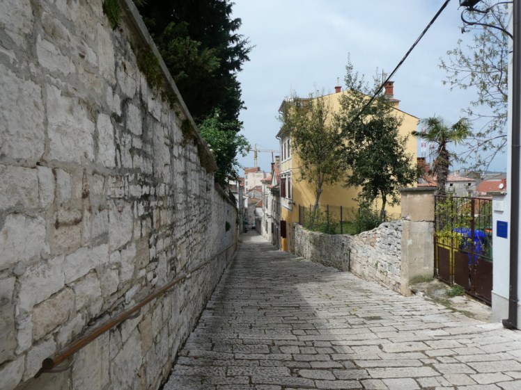 A cobbled street sloping steeply downwards away from the steps leaving the fort. On one side is a high stone wall, on the other a low wall enclosing a garden and then some houses, one painted yellow.
