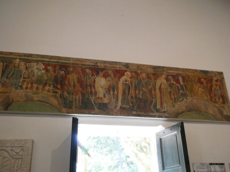A large fresco above the church's double doors, mostly in shades of red, showing a procession of people accompanied by skeletons.