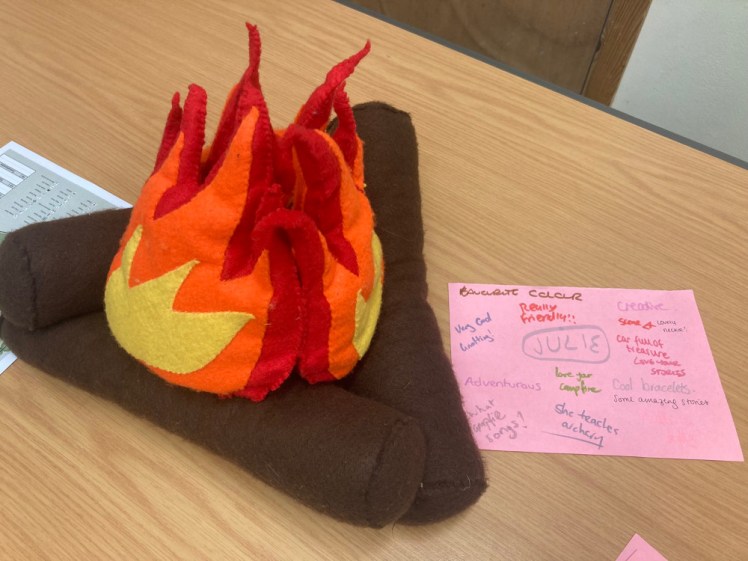 A felt campfire on a table next to a piece of paper with my name in the middle and positive comments all around it in different coloured pens and different handwriting.