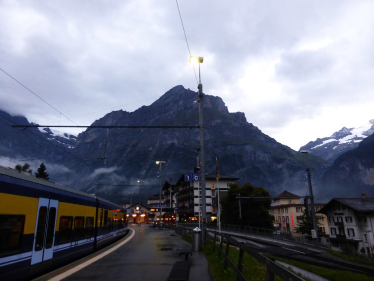 Grindelwald in the evening. The town looks dark but it's not really - the sky is still quite light but the mountains surrounding the town block out a lot of the light. The photo is on the station platform with a navy and yellow train standing there on my left.