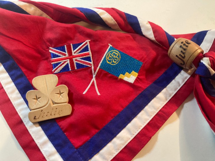 My red international neckerchief with blue and white borders, lying on my desk with a small carved wooden Trefoil-shaped badge lying on it. The neckerchief has a wooden woggle like an overgrown bead threaded onto it. Both have my name burned into them.