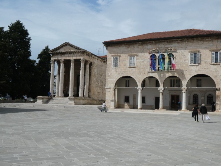 A view across the Forum, featuring the Temple of Augustus, a small but reasonably tall columned temple on the left and a large medieval City Hall on the right, with a smooth paved square in front of them.