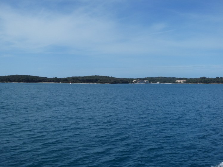 Out at sea, the Brijuni islands on the horizon. The sea is very blue - a vivid teal under the blue sky and the tree-covered island forms a barrier keeping the two apart.