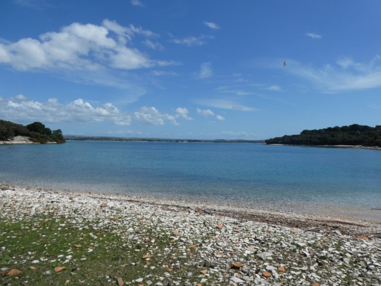 A turquoise-blue bay, longer than it is wide, looking from the shingle shore towards the entrance to the open sea, with the mainland visible on the horizon beyond.