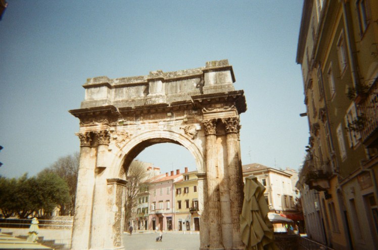 The Arch of the Sergii, a Roman arch looking dirty and aged but nonetheless glowing in the sun.