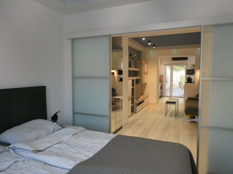In the foreground is the bedroom, with a grey cover over the foot of the bed and frosted glass sliding doors showing a view through the apartment to the back door. Everything is very tasteful and neutral except that the ceiling is a mid-blue instead of the expected white.