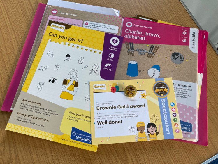 A folder of Brownie activity cards. On the left is a yellow Unit Meeting Activity card with a purple corner called Can You Get It?, on the right is a pink Skills Builder card and on top of them is a Brownie Gold Award certificate and pin badge - not to be awarded to the girl that ran the activity actually, that came next week.