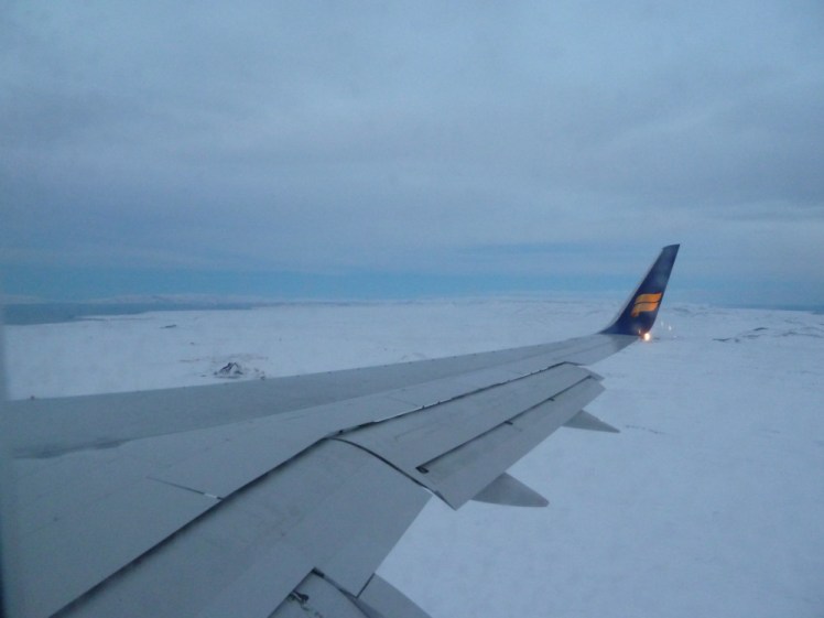 The view from the right-hand side of an Icelandair plane, as it approaches Keflavik. The sky is cloudy with hints of blue and the entire landscape is just snow.