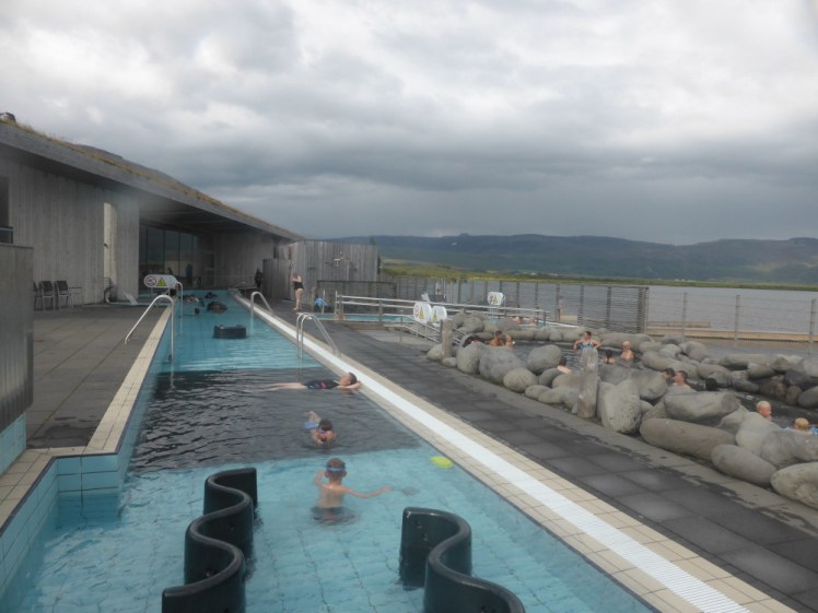 The complex at Laugarvatn Fontana, as seen from the raised hot pot. Most of the photo is dominated by a long shallow tiled pool. To the right, behind the boulders, is a hotter pool with a more natural bottom and behind that is a small swimming pool. You can also go out through the gate and swim in the lake behind the fence if you really want to.