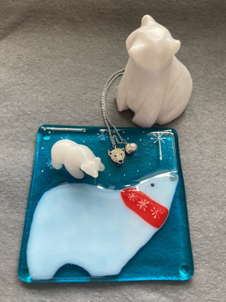 A glass coaster with a polar bear looking up at a starry sky. On top of it is a small soapstone polar bear and my Swarovski polar bear necklace. Next to it, with the chain of the necklace disappearing behind it, is a more stylised soapstone polar sitting up on its haunches.
