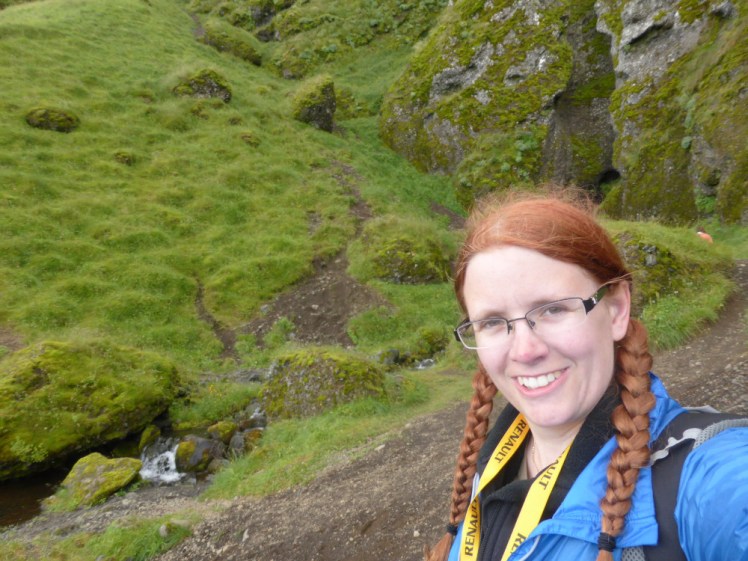 A selfie from 2014 at Rauðfeldsgjá, with the plaits and some really bright ginger-red hair. Rauðfeldsgjá is a tiny crack in the green-covered mountain behind me, barely visible.