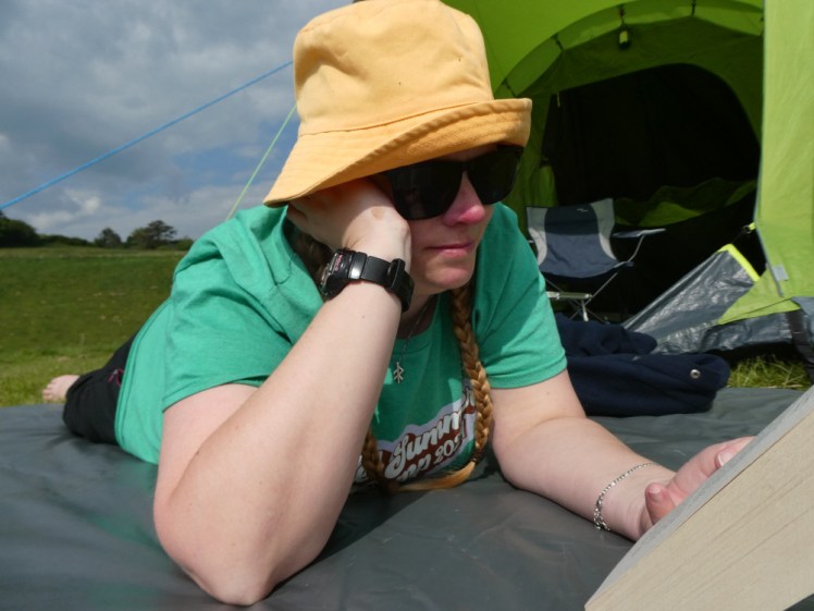Me, in a green t-shirt and yellow bucket hat, lying on a groundsheet outside my tent, reading.