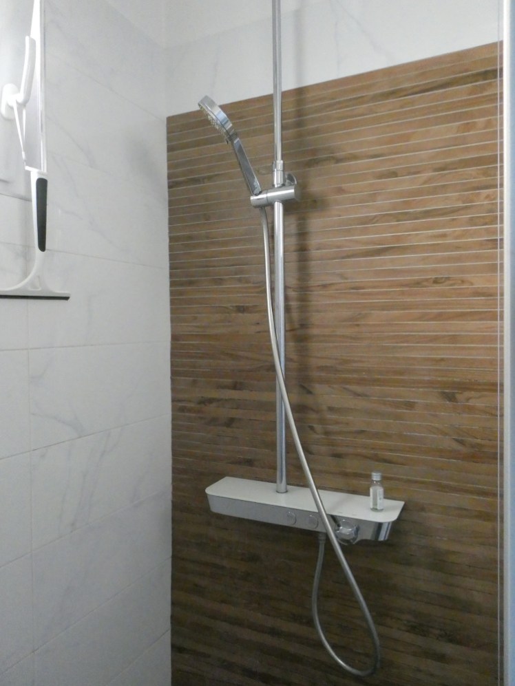 The shower. The control unit is set into a little thick shelf and consists of two buttons set right into the shelf and a temperature dial. The shower is mostly tiled in marble-effect white tiles but the shower is on a made to look like very narrow wooden slats.