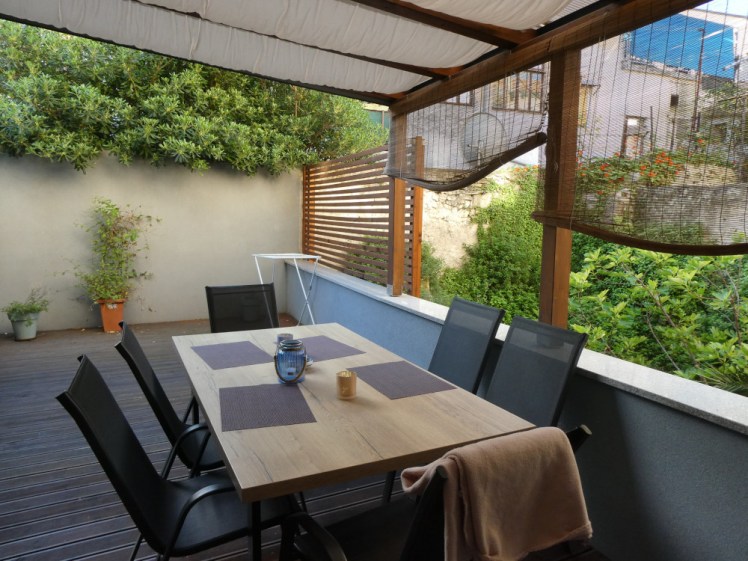 A table with placemats and six chairs set out on a terrace. Above the table is a wooden structure covered with plastic and then hung on the inside with white cloth to keep off both sun and rain. At the far end of the terrace, it's open to the sky. There's vegetation growing on the back wall and also in the yard next to and below the terrace, reaching up to above the terrace's level.