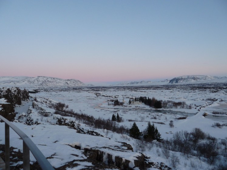 Þingvellir National Park as seen from the viewpoint in winter. The sky is pink on the horizon, turning light blue up higher and the lava field below the cliffs is just white with snow and with fir trees poking out all over the place.