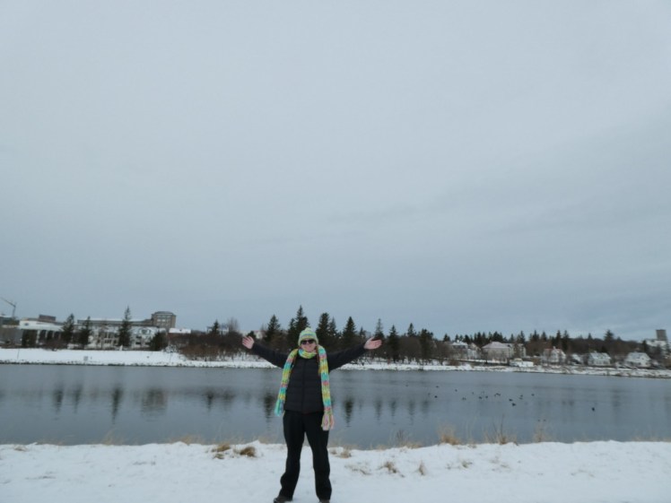 A timer selfie by the edge of a lake in Iceland. The sky is grey with incoming snow, the lake is grey, the banks of the lake are white with snow and I'm wearing a long scarf and matching hat in yarn that's somehow neon bright and pastel at the same time.