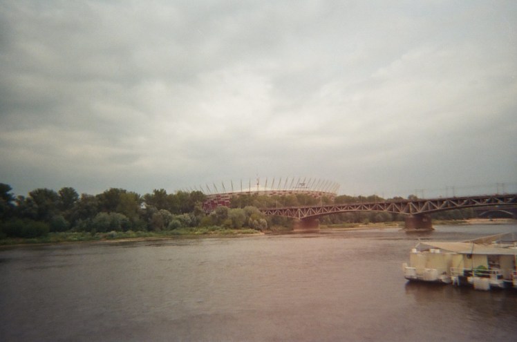 Warsaw National Stadium across the river, a kind of red wicker basket looming above the city.