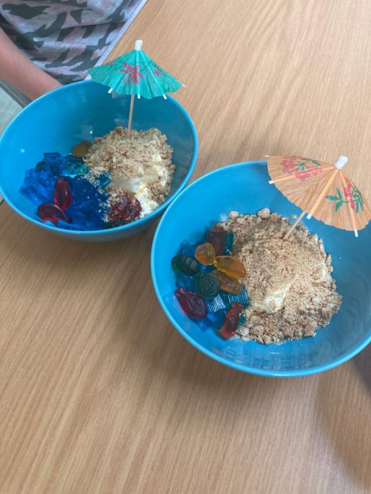 Two blue bowls containing beach scenes made of blue jelly with sea creature sweets and a beach made of ice cream with crumbled biscuit sand, plus paper cocktail umbrellas.