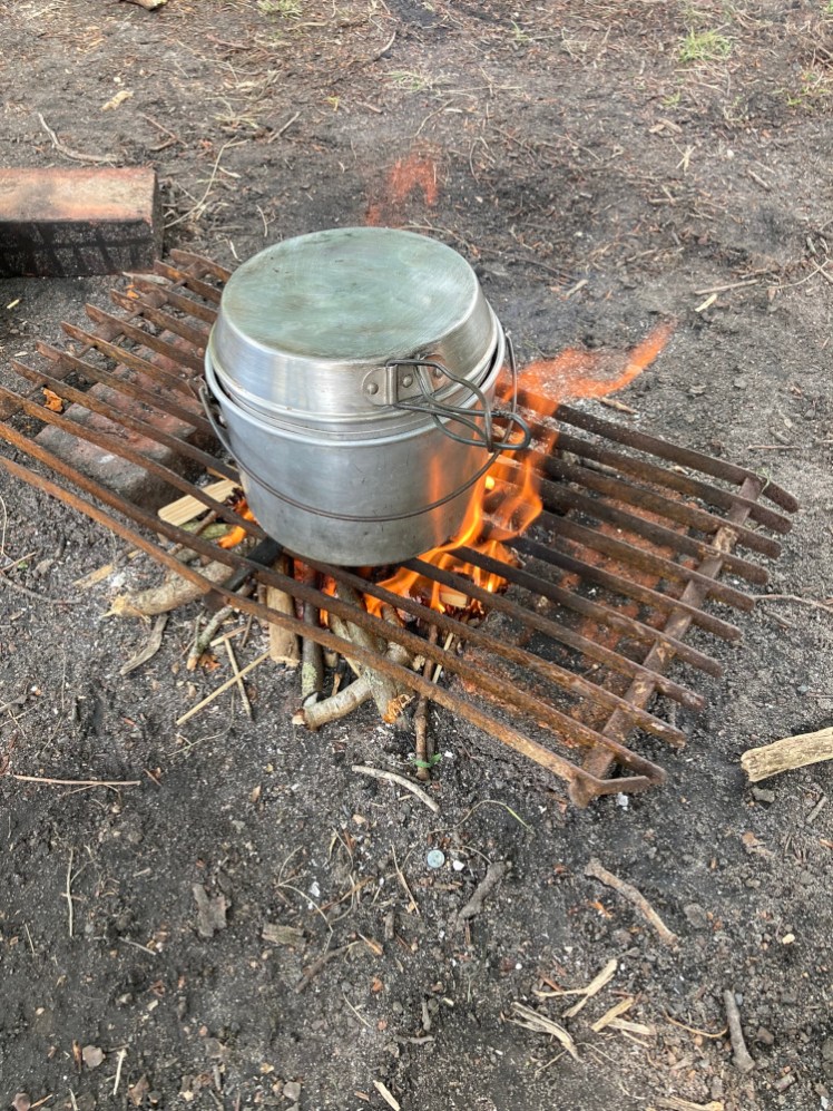 A billy, a lightweight aluminium cooking pot with a wire handle and matching lid, sitting on a grid over a fire. The pot is full of water and later it'll cook pasta.