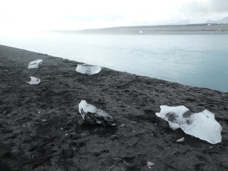A few small chunks of ice lying on a beach made of black sand, with an overexposed river flowing past behind them. This is the famous Diamond Beach, where the "diamond" content varies hugely from that advertised by social media.