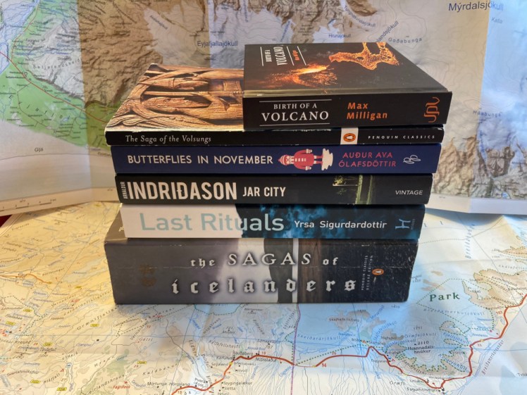 A pile of Icelandic books, including sagas, crime and a mini picture book of the 2021 eruption, all in a neat pile on top of an Iceland map with another map as the background.