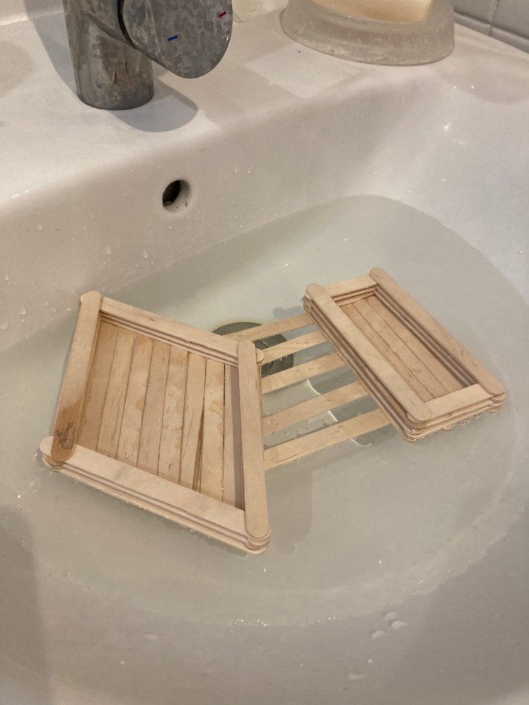 A raft made out of lolly sticks floating in the sink. The main part is diamond-shaped, the smaller outrigger is a thin rectangle and the two are joined by five sticks to create a very stable little boat.
