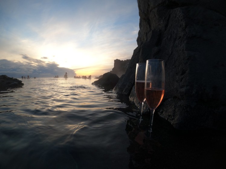 The Sky Lagoon at sunset, with an explosion of pastel blues, pinks and yellows reflecting on the water. In the foreground, two glasses of pink wine are almost silhouetted against the cliff edge that forms the inside of the lagoon.