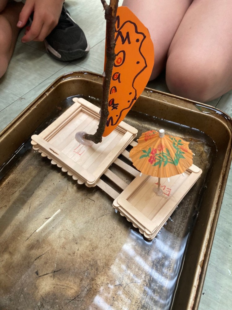 A slightly grubby roasting pan full of water, in which a boat made out of lolly sticks is floating. The boat has a mast made from a twig, an orange sail with Moana written down it and the umbrella from the sundae is glued into the outrigger. In the background: a pair of Brownie knees watching her boat sail.