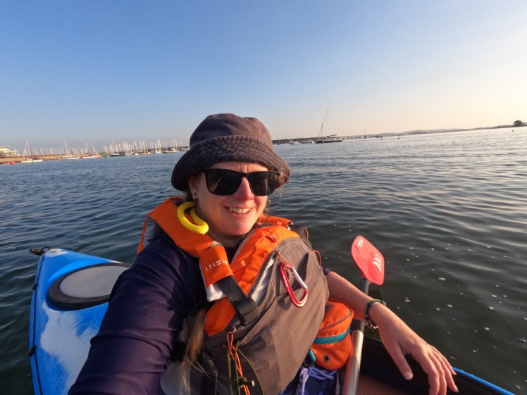 A selfie in a kayak out at sea at the boathouse. I'm wearing a stained navy bucket hat, sunglasses and an orange and grey buoyancy aid and piloting a big blue kayak.