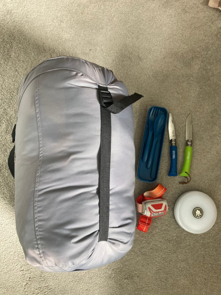 My camping stuff: sleeping bag in a grey stuffsack, little plastic box of plastic cutlery, two sharp knives, a headtorch and a circular tent light.