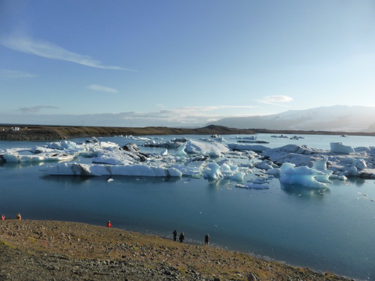 Jökulsárló as seen from a hillock. The water is vast and flat and very blue and there are chunks of ice floating around in it - icebergs of various colours, decorations, shapes and sizes.
