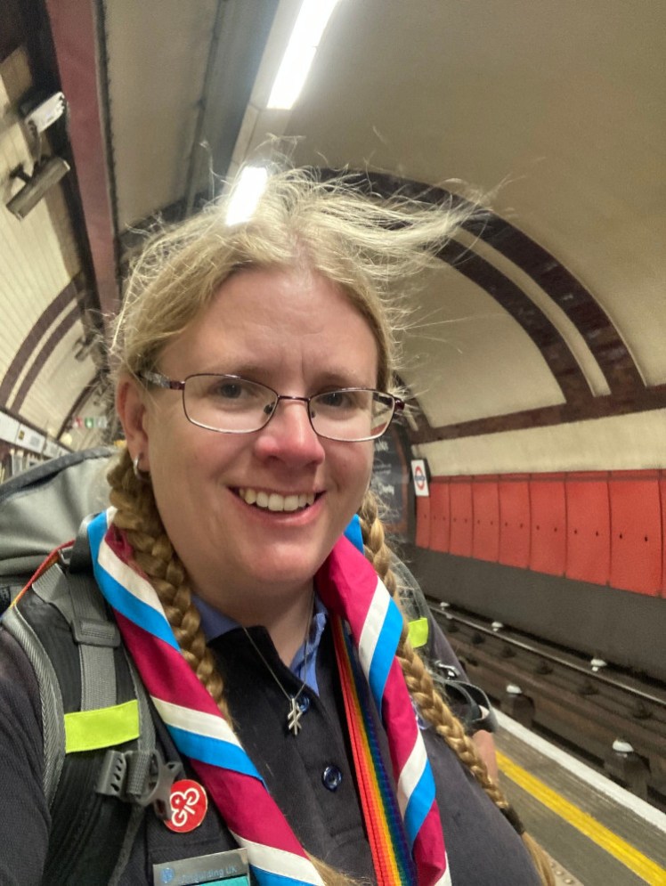 Me on a Tube platform, my hair a little blown by the wind underground. I'm wearing a pink, blue and white Region neckerchief, my Guide uniform and a backpack.
