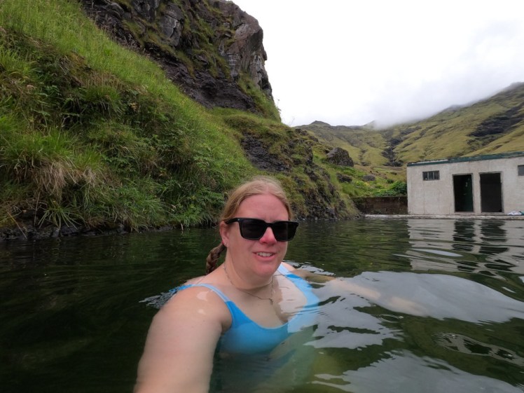 A selfie in the slightly green, slightly opaque water of Seljavallalaug. The green-covered mountain behind it forms the back wall, there's a little white hut for changing and behind that are more green mountains.
