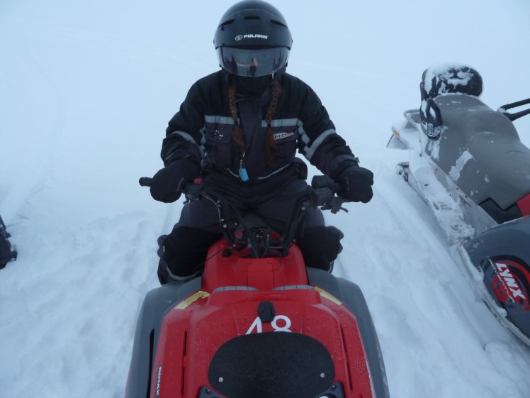 Me in a black snowsuit and black helmet with black visor, sitting on a red snowmobilie on a background of snow.