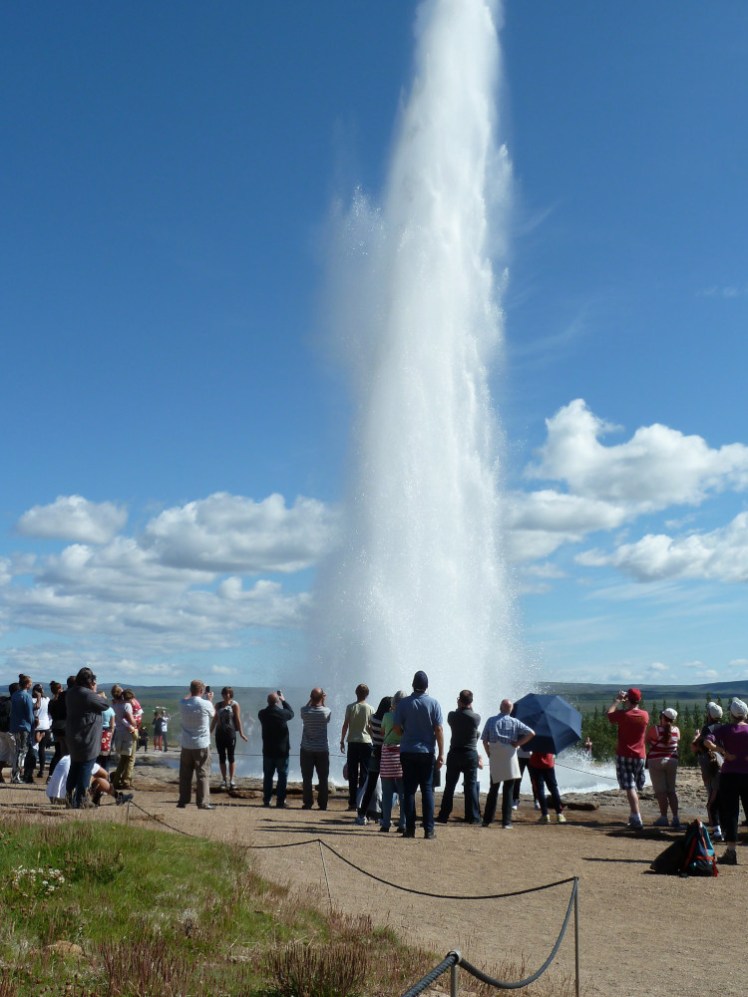 Strokkur erupting so far it goes off the top of the vertical photo. Lots of tourists are gathered in a circle around the geysir.