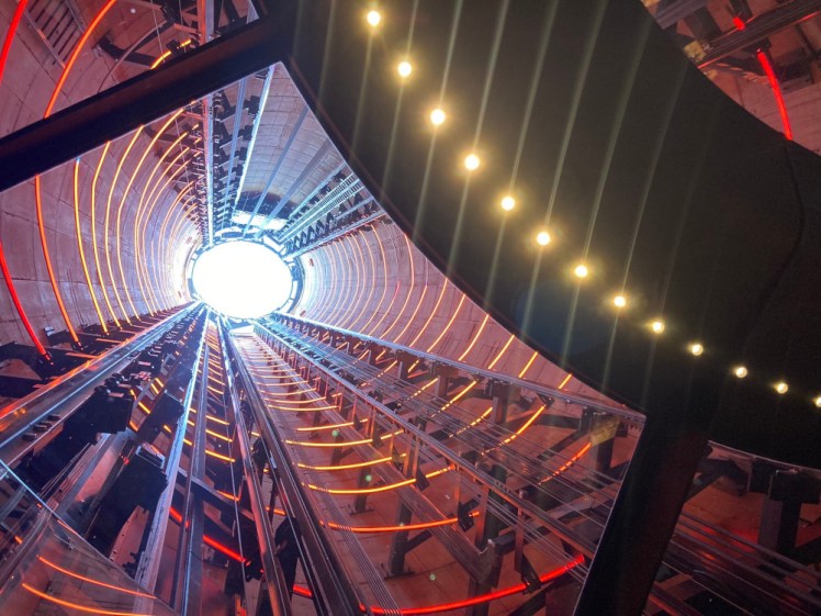 Looking up the chimney. The ceiling of the lift is encircled by little LEDs which are all starring spectacularly. Above it, the chimney interior is wrapped in circles of red, orange and yellow lights, with the lift metalwork almost blue from the light coming down the top of the chimney.
