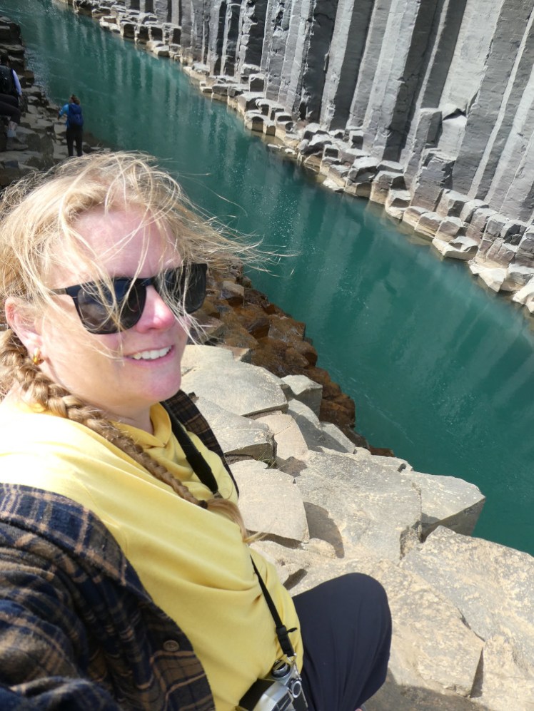A selfie at Stuðlagil, with a narrow ribbon of green-blue water, perfect straight basalt columns going right down to the river and me, in a yellow top with my hair all over my face, sitting above it on a pile of hexagonal rocks.