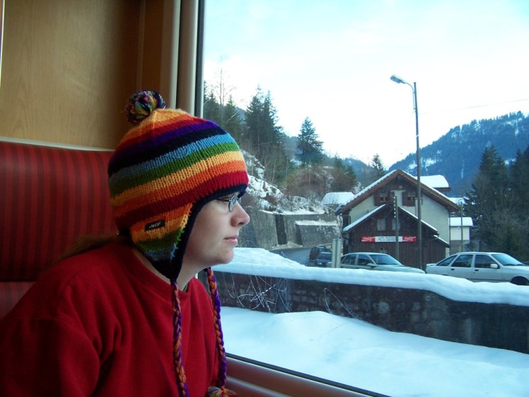 Me sitting on a train in Switzerland looking out at the snow, wearing a red jumper and a rainbow-striped hat with earflaps.