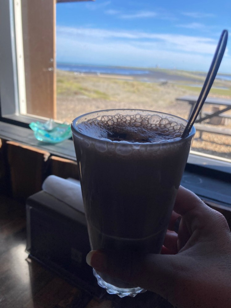Hot chocolate in a glass, next to a window looking out at the headland.