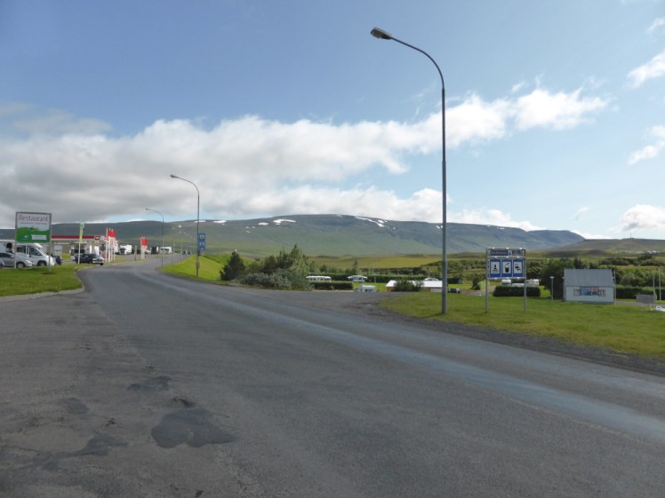 Blönduós as seen from the side of the Ring Road. There's a fuel station with red flags up the road on the left but the entire right side is rolling hills and a campsite with hot tubs and mountains in the background. It looks pretty idyllic as towns go.